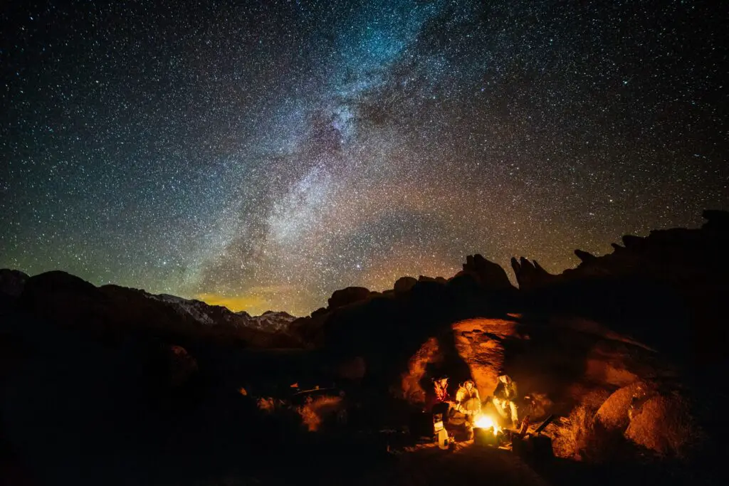 A sky full of stars over a mountain backdrop with a family enjoying a campfire together.
